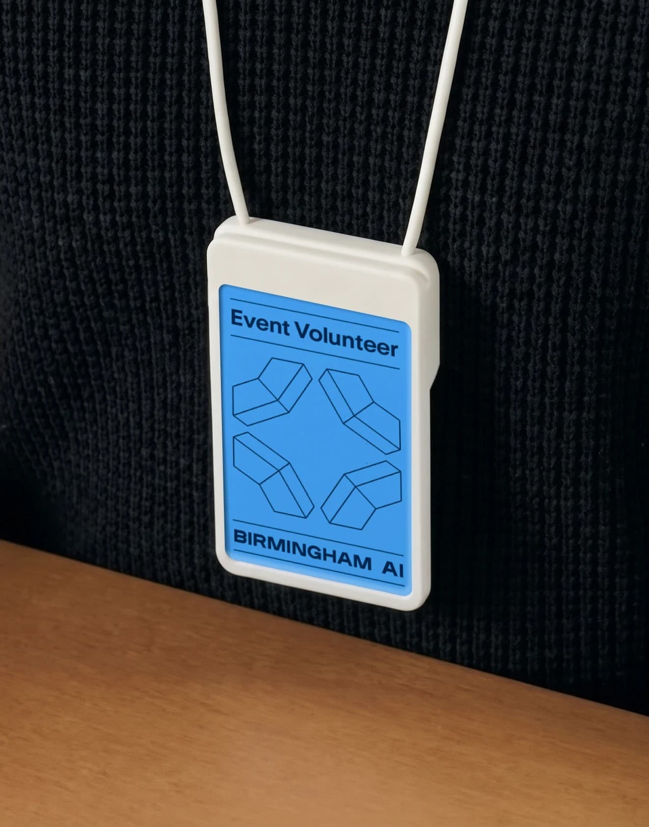 A Birmingham AI Event Volunteer badge on a white lanyard, the blue card with the wireframe mark worn against a dark knit sweater above a wooden table