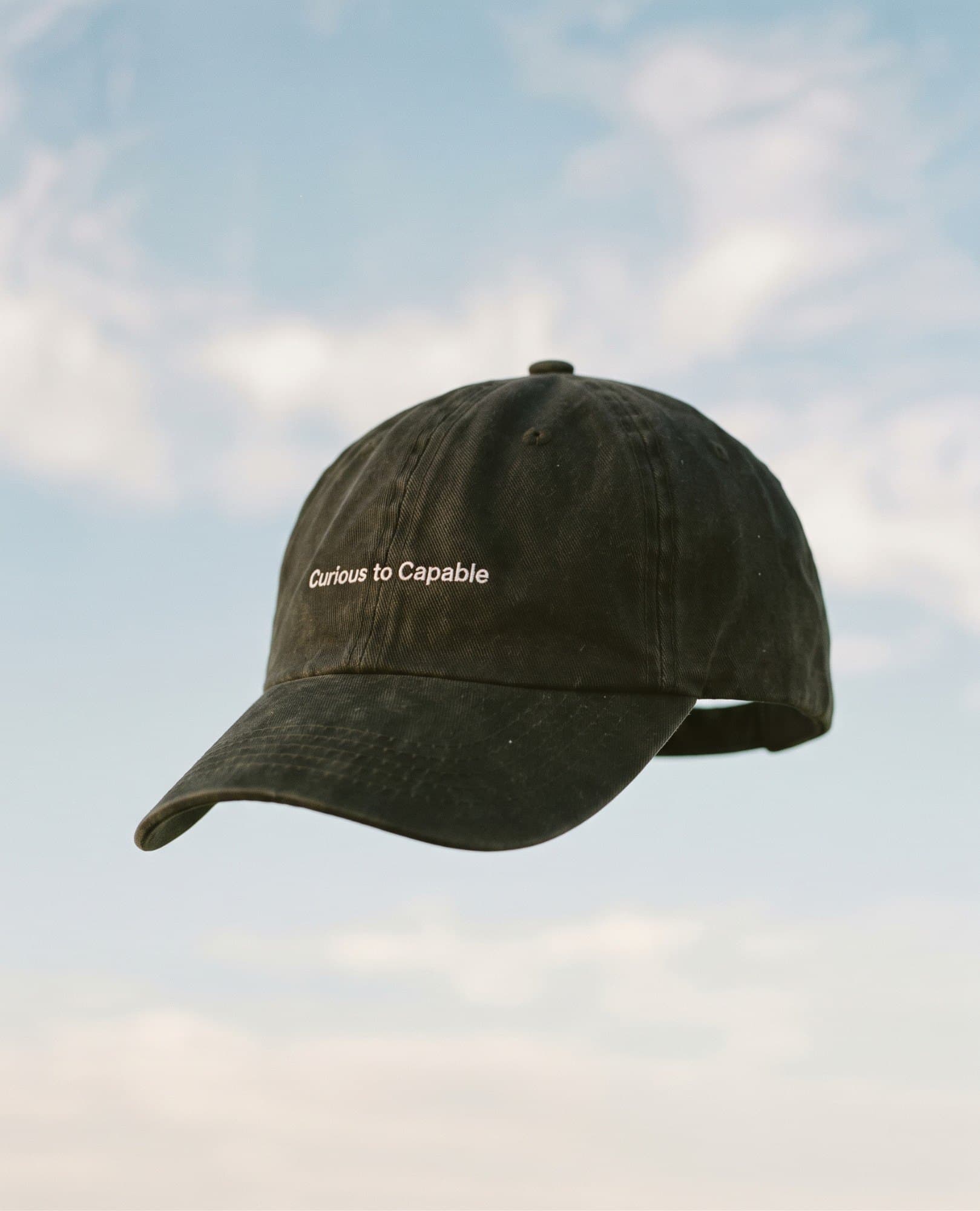A washed black cap embroidered Curious to Capable, floating against a blue sky